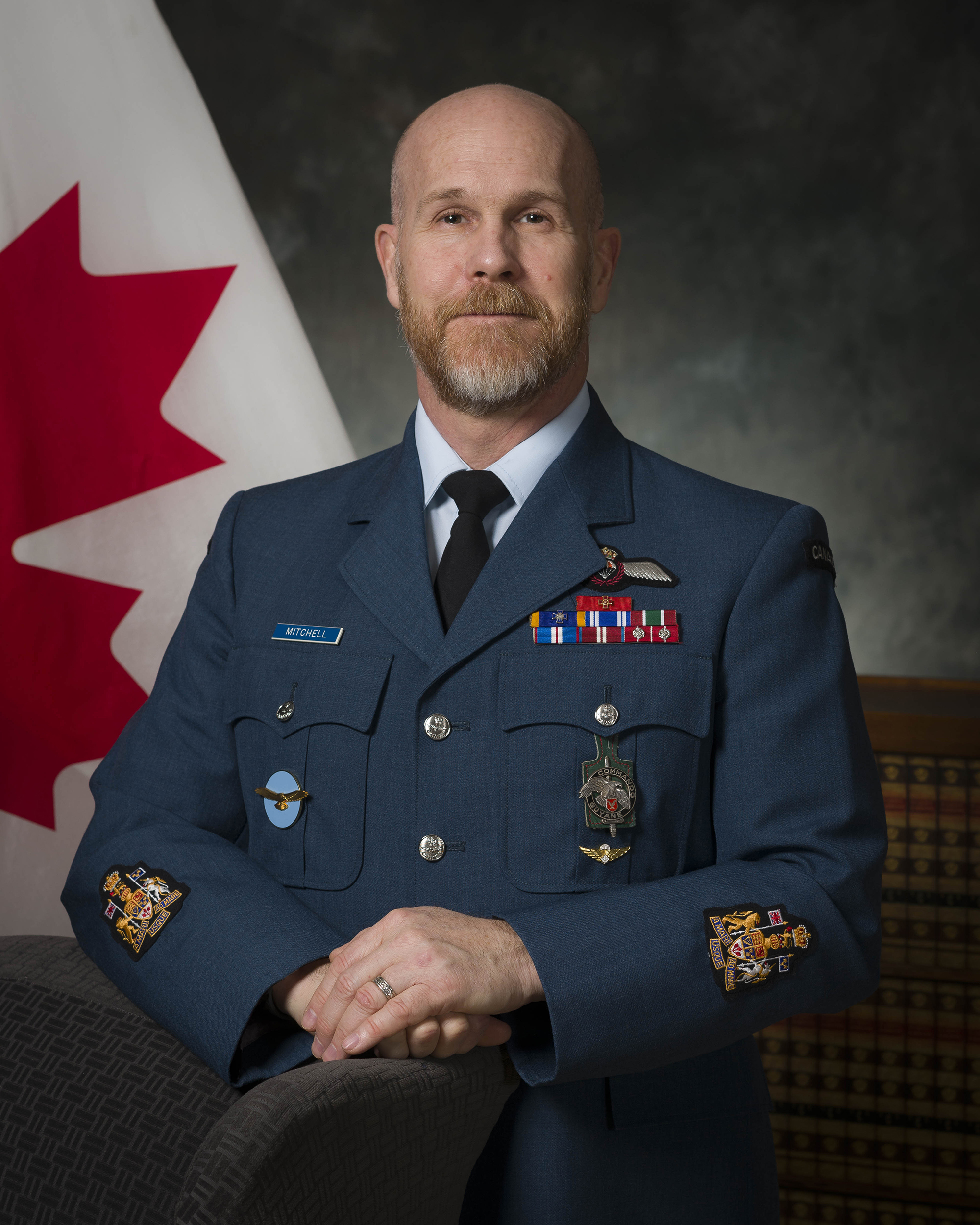 A photo of a man wearing a blue military uniform with medals. Behind him is a Canadian flag.