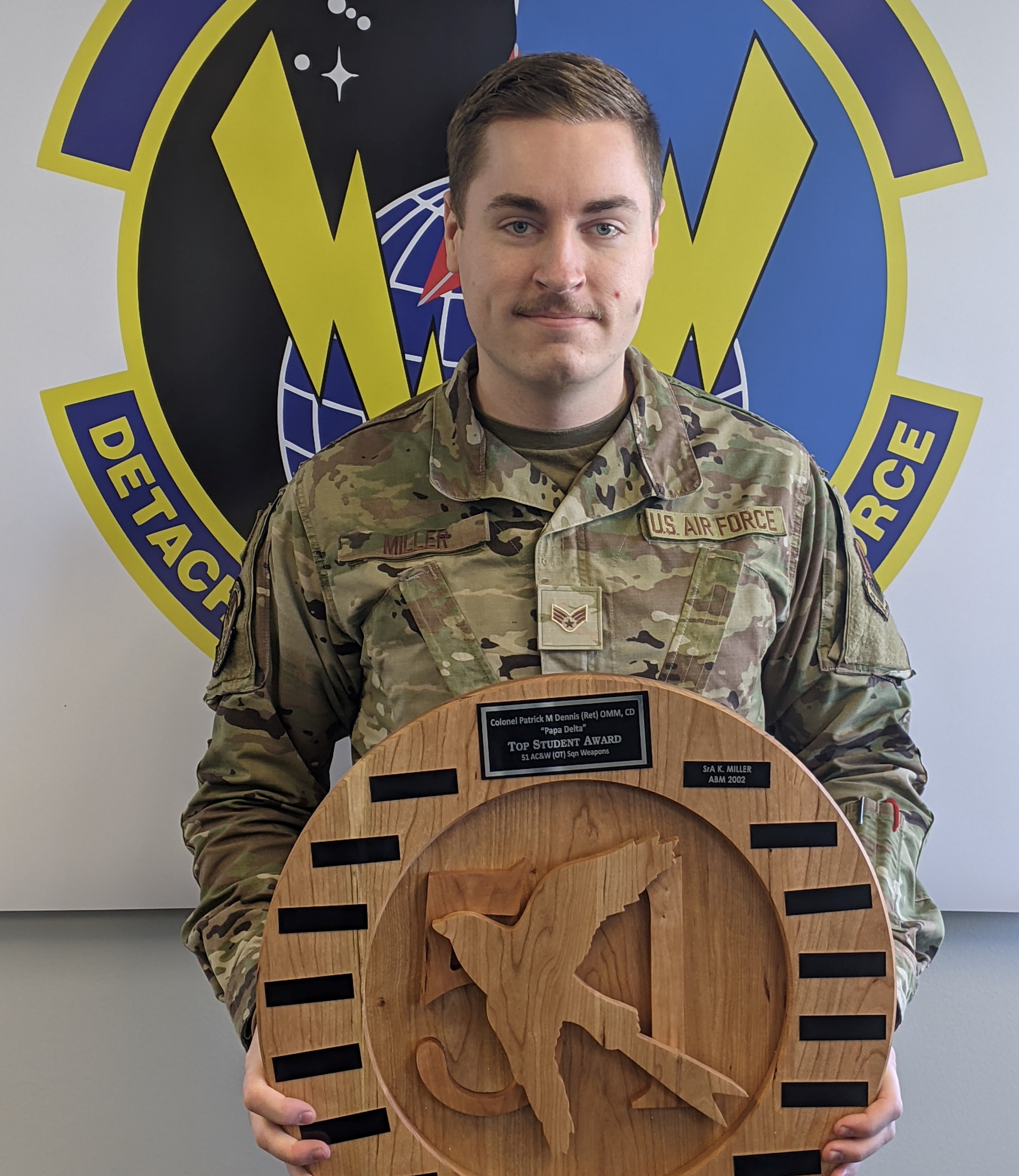 Un homme souriant vêtu d’un uniforme à camouflage tient un prix rond en bois. Sur le mur derrière lui, on voit un gros insigne militaire.