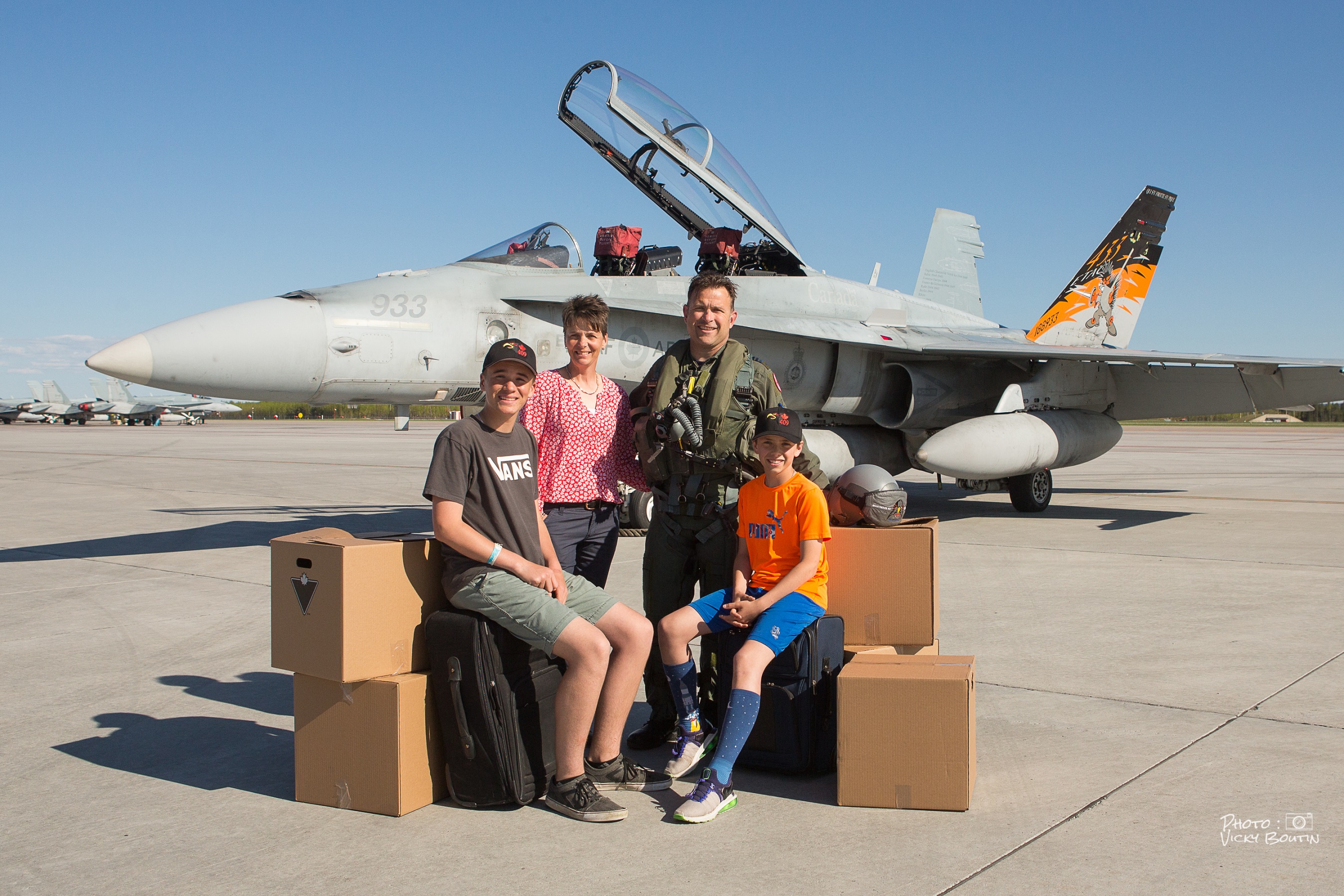 Two adults standing, one wearing a military flight suit, and two children sitting on luggage among cardboard boxes. Behind them is a grey jet fighter with its canopy open. 