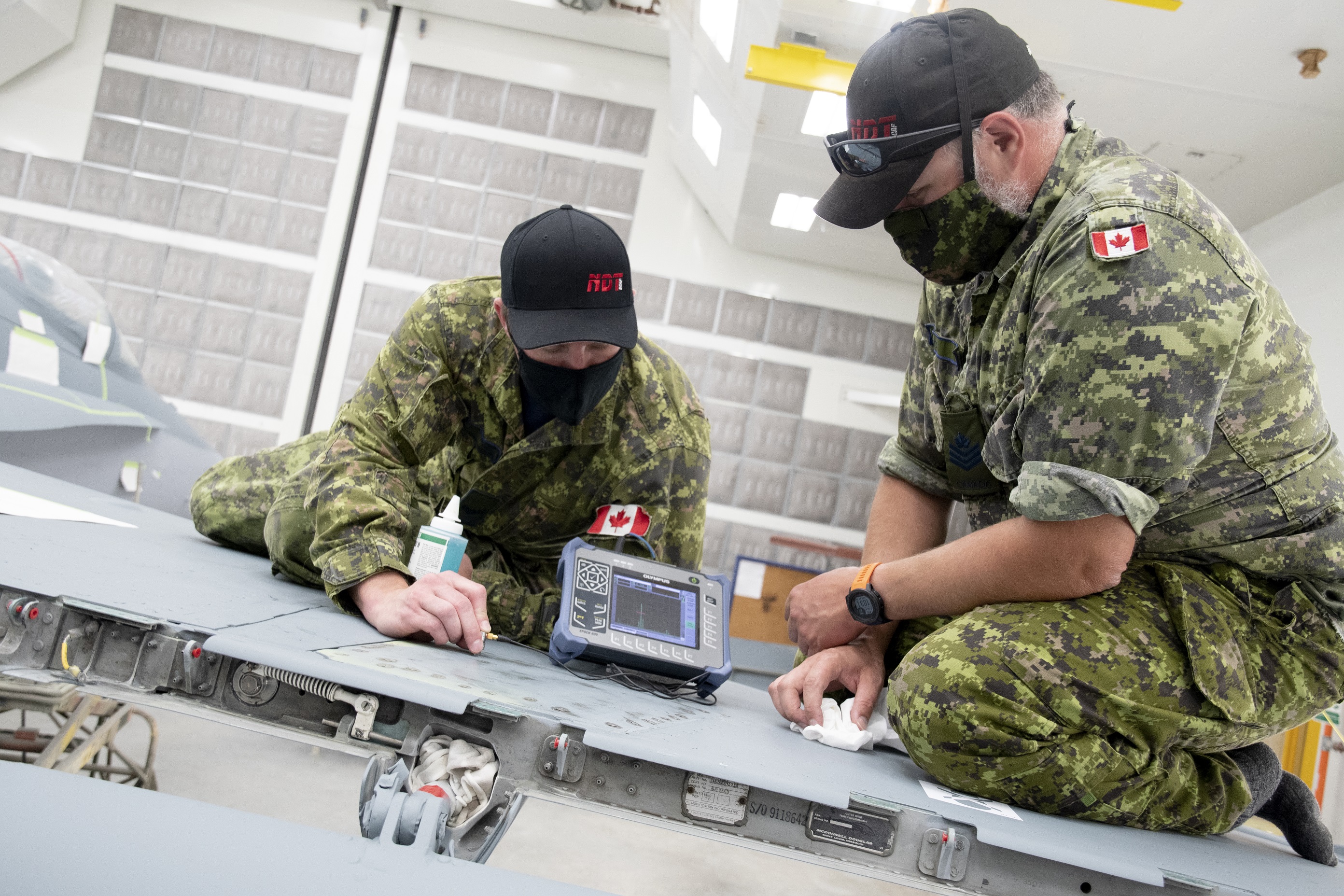 Sergeant Adam Smith (right), NDT Development Supervisor at Trenton-based ATESS, watches the screen on a portable ultrasonic device while 8 AMS NDT Support technician Corporal Eddy Ford (left) uses pulse echo ultrasonic techniques to inspect the skin (surface) of a CF-18 Hornet’s wing for delaminations. 