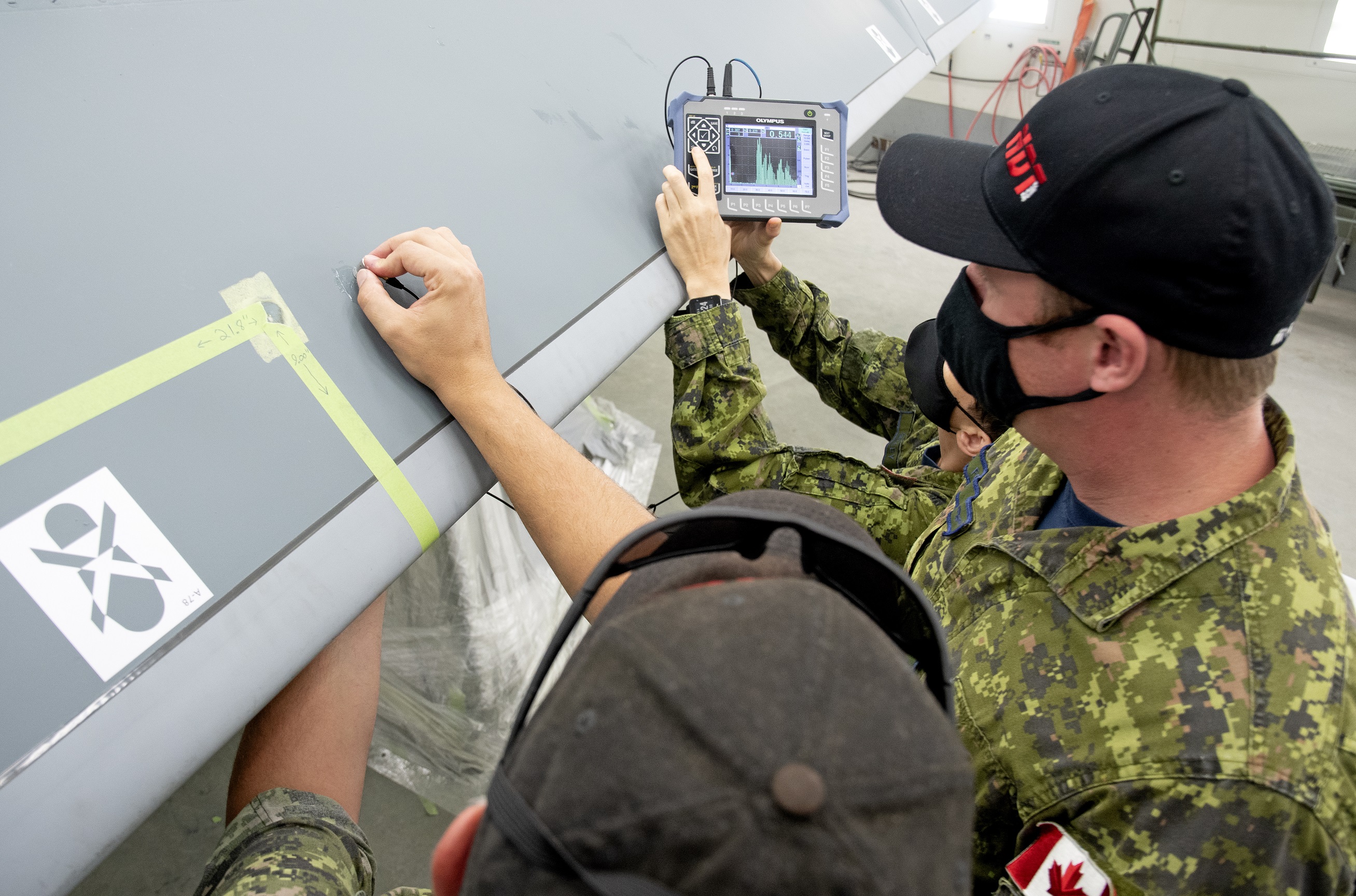 ATESS NDT Development Supervisor Sergeant Adam Smith (left) holds a sensor on the leading edge flap of a CF-18 Hornet while 8 AMS (air maintenance squadron) NDT Support Technician Corporal Eddy Ford (centre) and Cpl Maggie Selley (left) of ATESS’ NDT Training and Development use ultrasonic transmissions to look for signs of damage from the skin (surface) of the aircraft to the structures and spaces underneath the surface.  