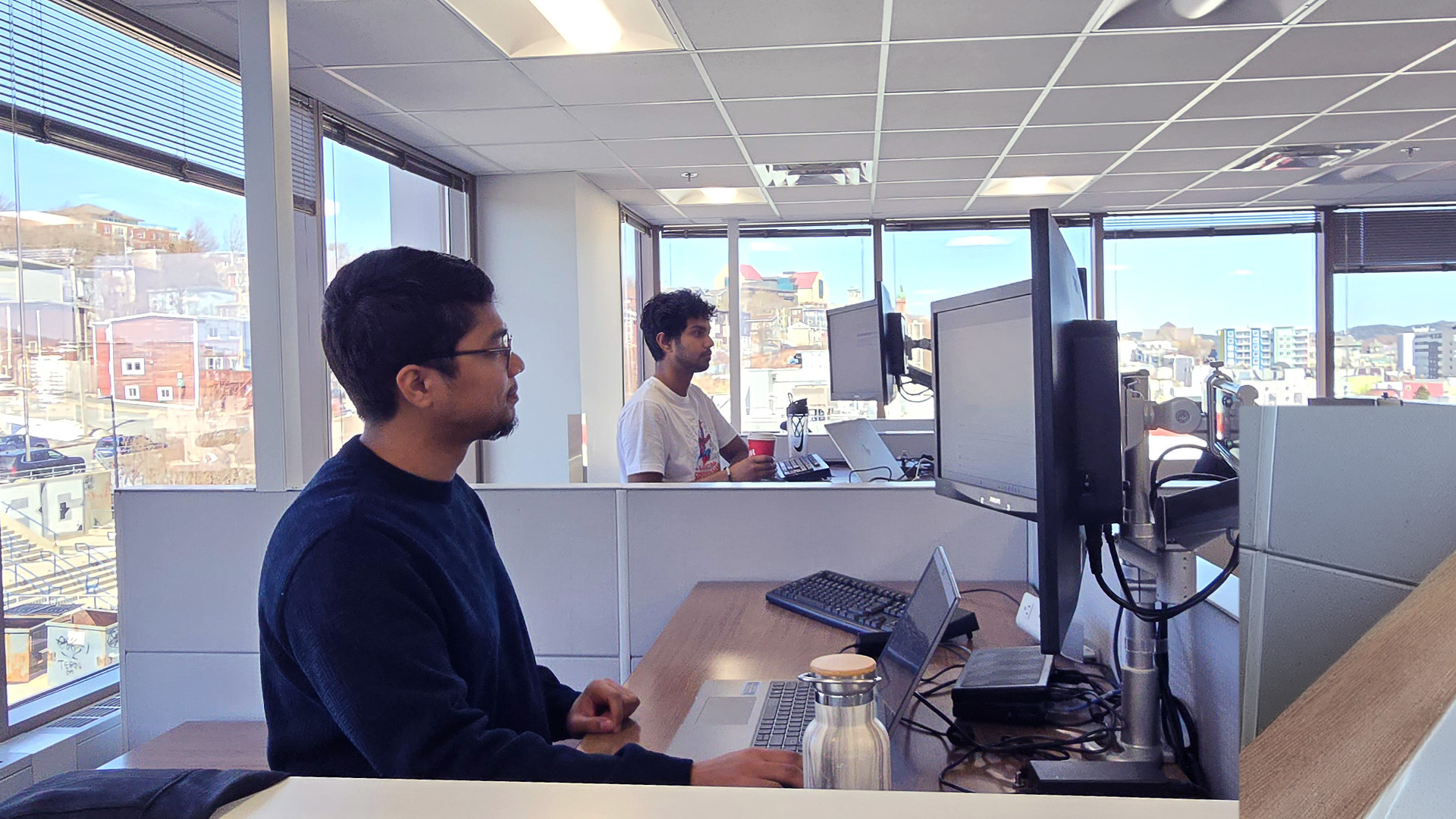 Two students from the CanAI Garage Program work at standing desks in an office. In the background, windows show views of St. John’s, Newfoundland and Labrador.