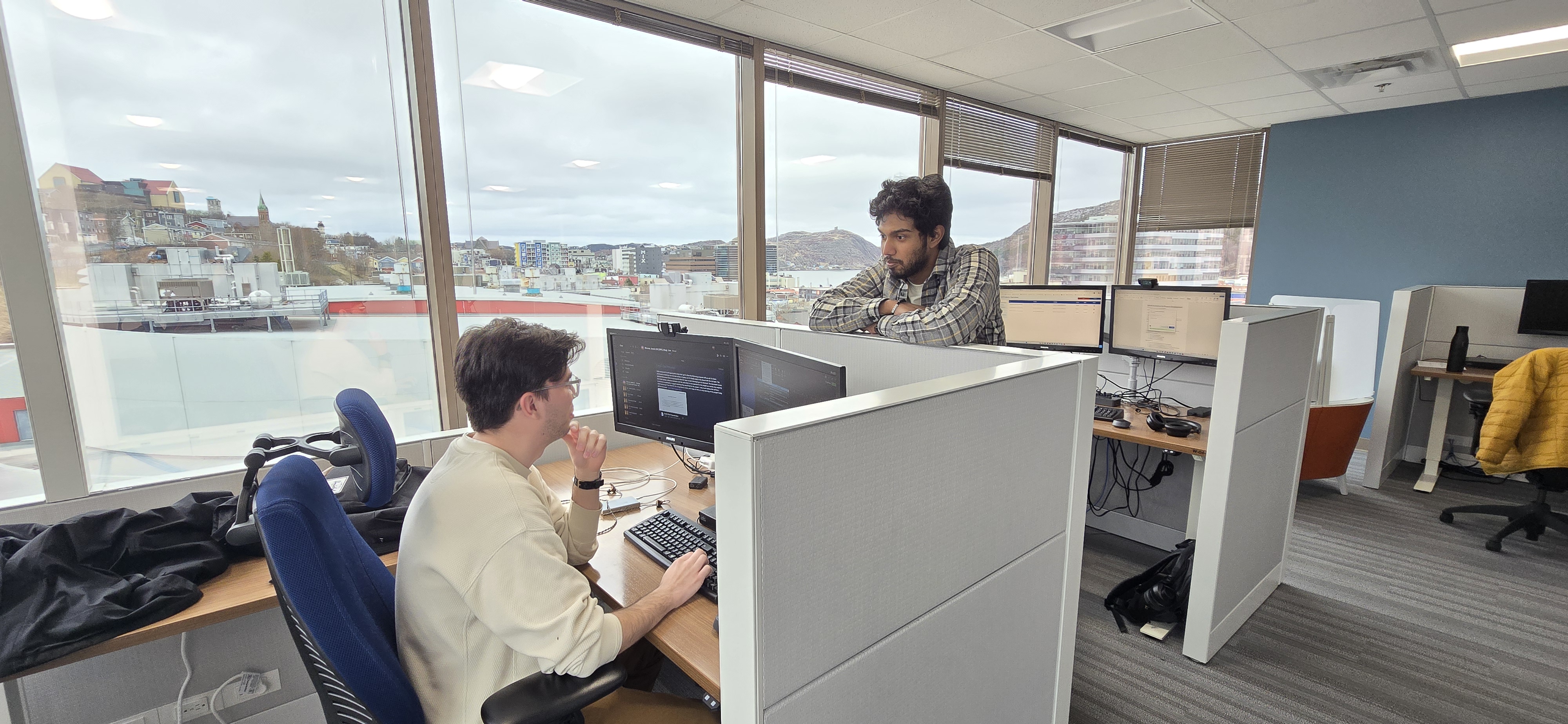 Two students from the CanAI Garage Program discuss a project over a cubicle wall. In the background, windows show views of St. John’s, Newfoundland and Labrador.