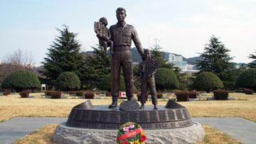 Life size sculpture of a soldier holding a baby and his left hand is on the shoulder of a young boy who is standing beside him