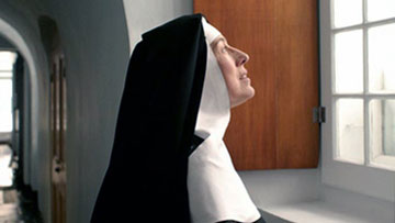A nun standing at a window looking out towards the sky