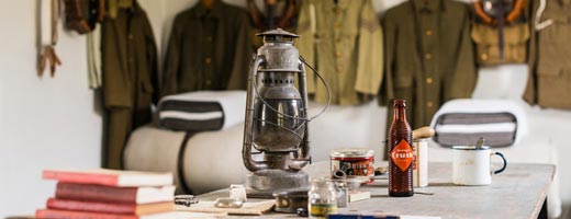 An oil lamp sits atop a table of books, bottles and jars. Green army coats are hanging in the background