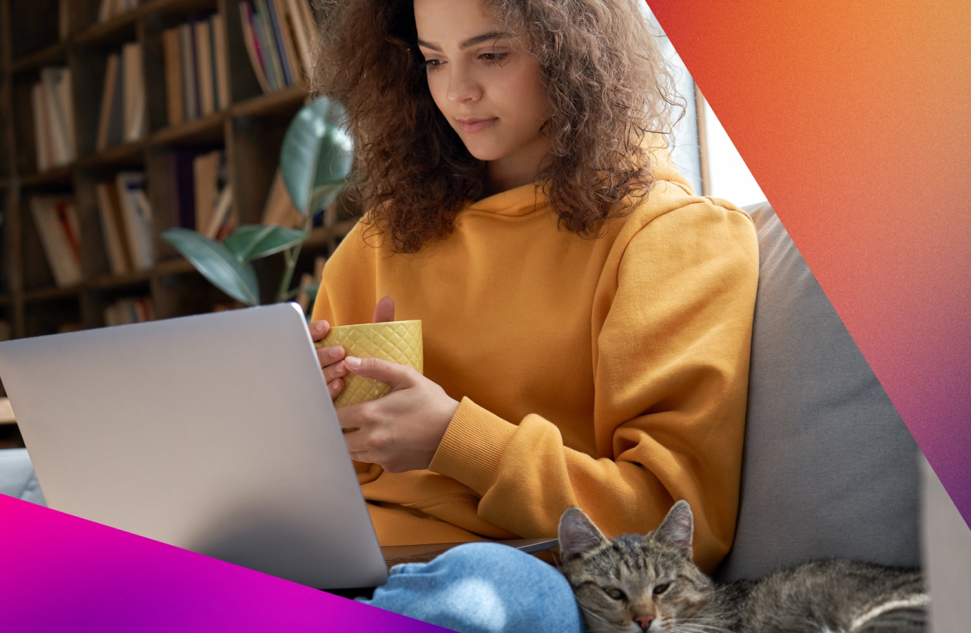 A woman sits on a couch with a laptop, holding a mug while a cat rests beside her.