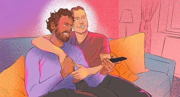Two men sitting together on a couch, smiling and holding hands, with one man's arm around the other.